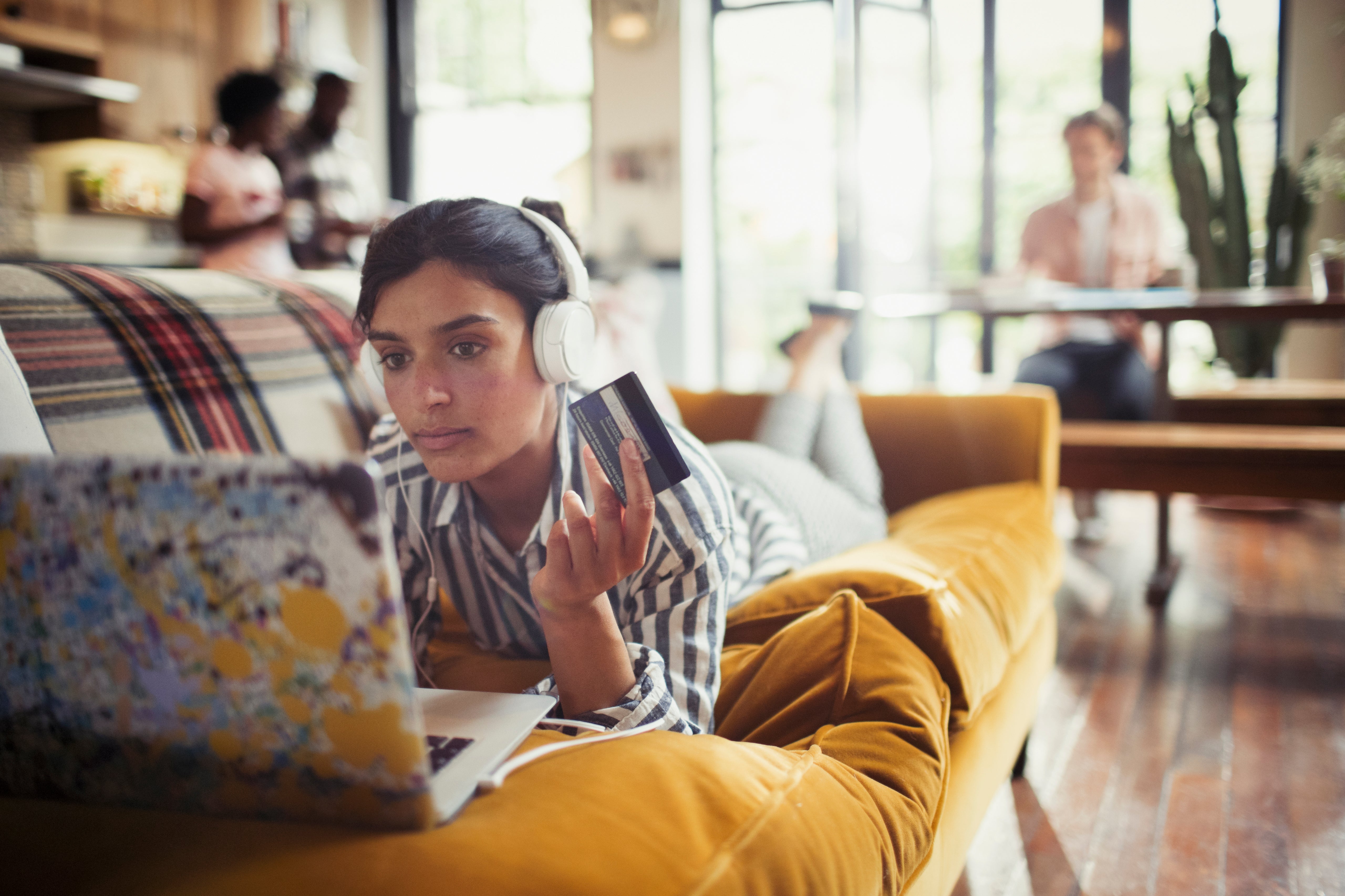 Young woman with headphones and credit card online shopping at laptop on living room sofa